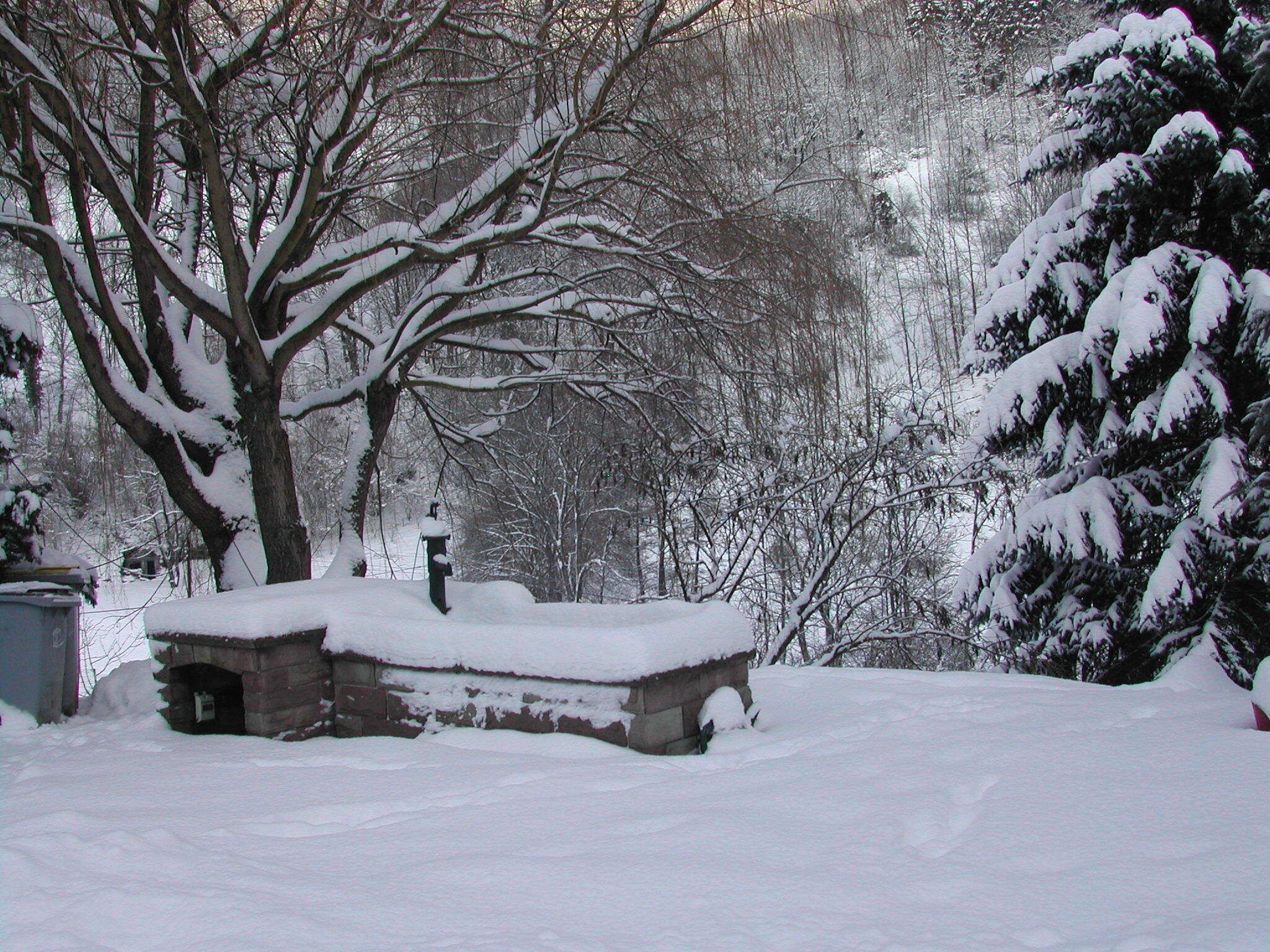 Ein verschneiter Außenbereich mit einer Steinsitzbank und einer kleinen Struktur, umgeben von Bäumen und schneebedeckten Ästen.