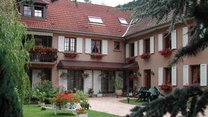 Un edificio de dos pisos con tejado de tejas rojas, contraventanas blancas y macetas con flores en las ventanas. Se aprecia un patio empedrado con un banco y plantas en macetas.
