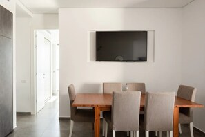Een eetgedeelte met een houten tafel en vier stoelen, een flatscreen televisie aan de muur en een deur naar een andere kamer.