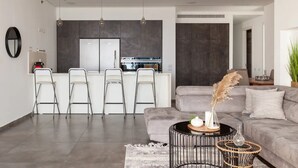 Een moderne woonkamer met een grijze bank, een salontafel en een eetgedeelte, met een keuken op de achtergrond.