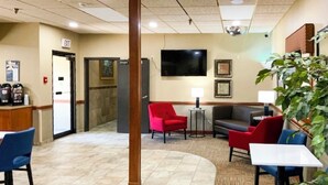 A lobby with a reception desk, a coffee station, seating area with red and grey chairs, a TV on the wall, and a plant. - Studio 6 Minot, ND (Minot)