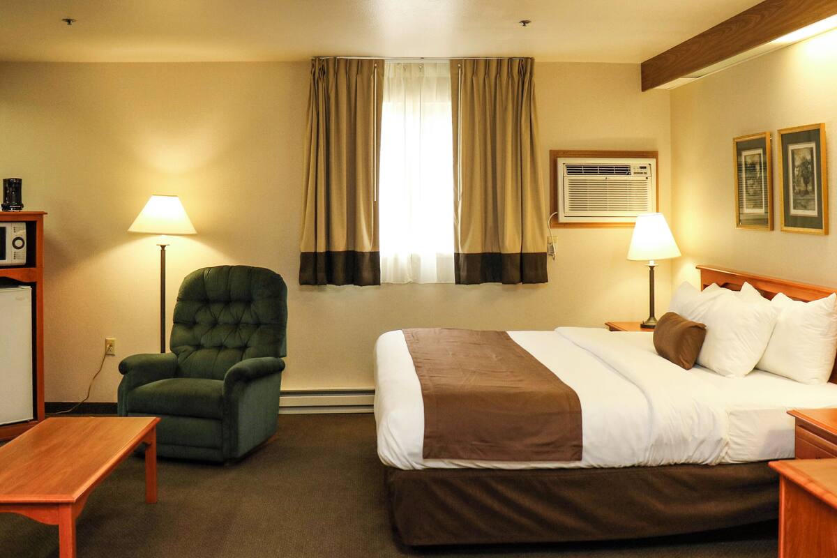 A hotel room with a bed, two bedside tables, a green armchair, a lamp, a window with curtains, an air conditioning unit, and framed pictures on the wall.