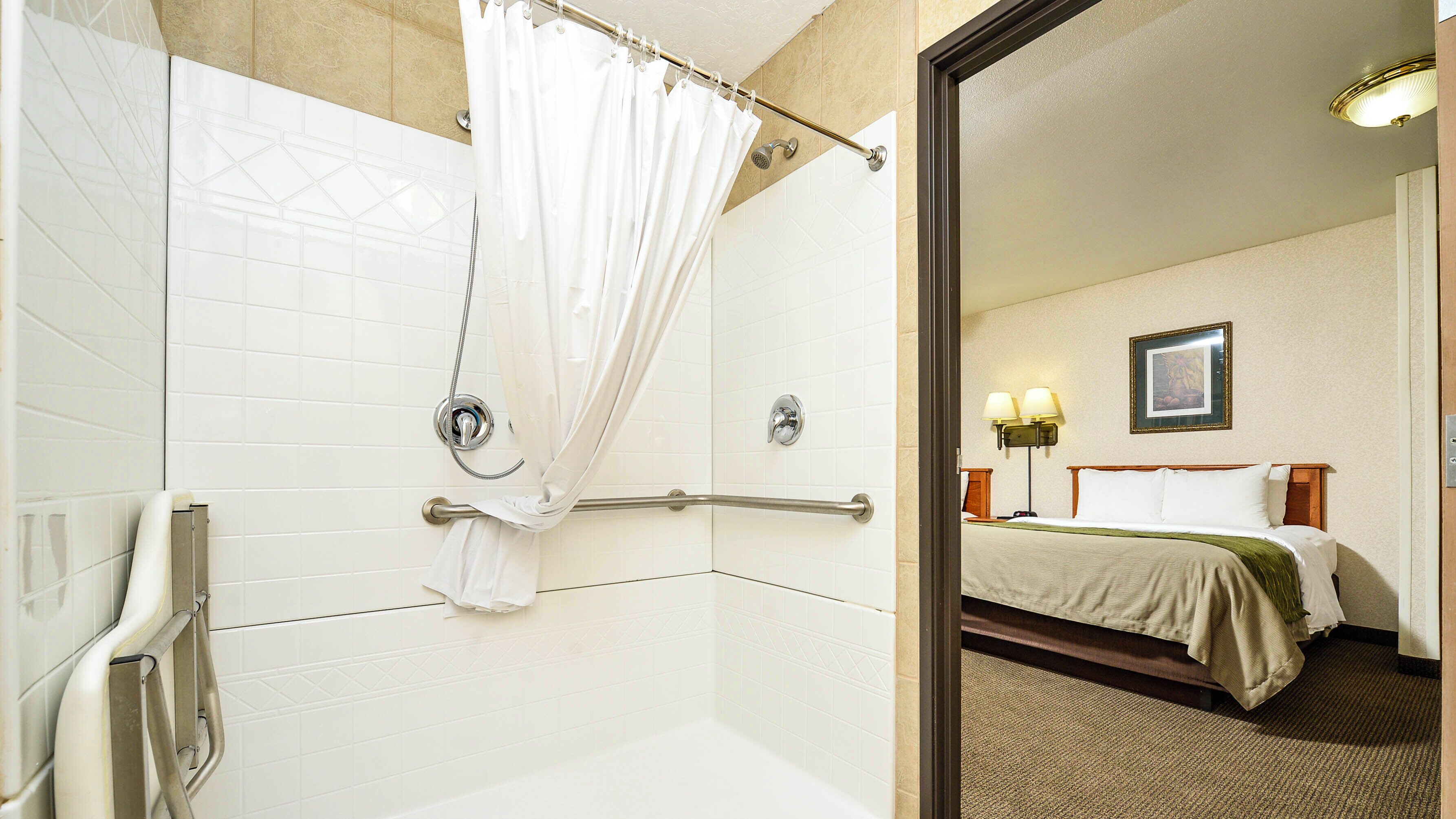 A hotel bathroom with a shower, a handrail, and a mirror reflecting a bedroom with a bed and a nightstand.