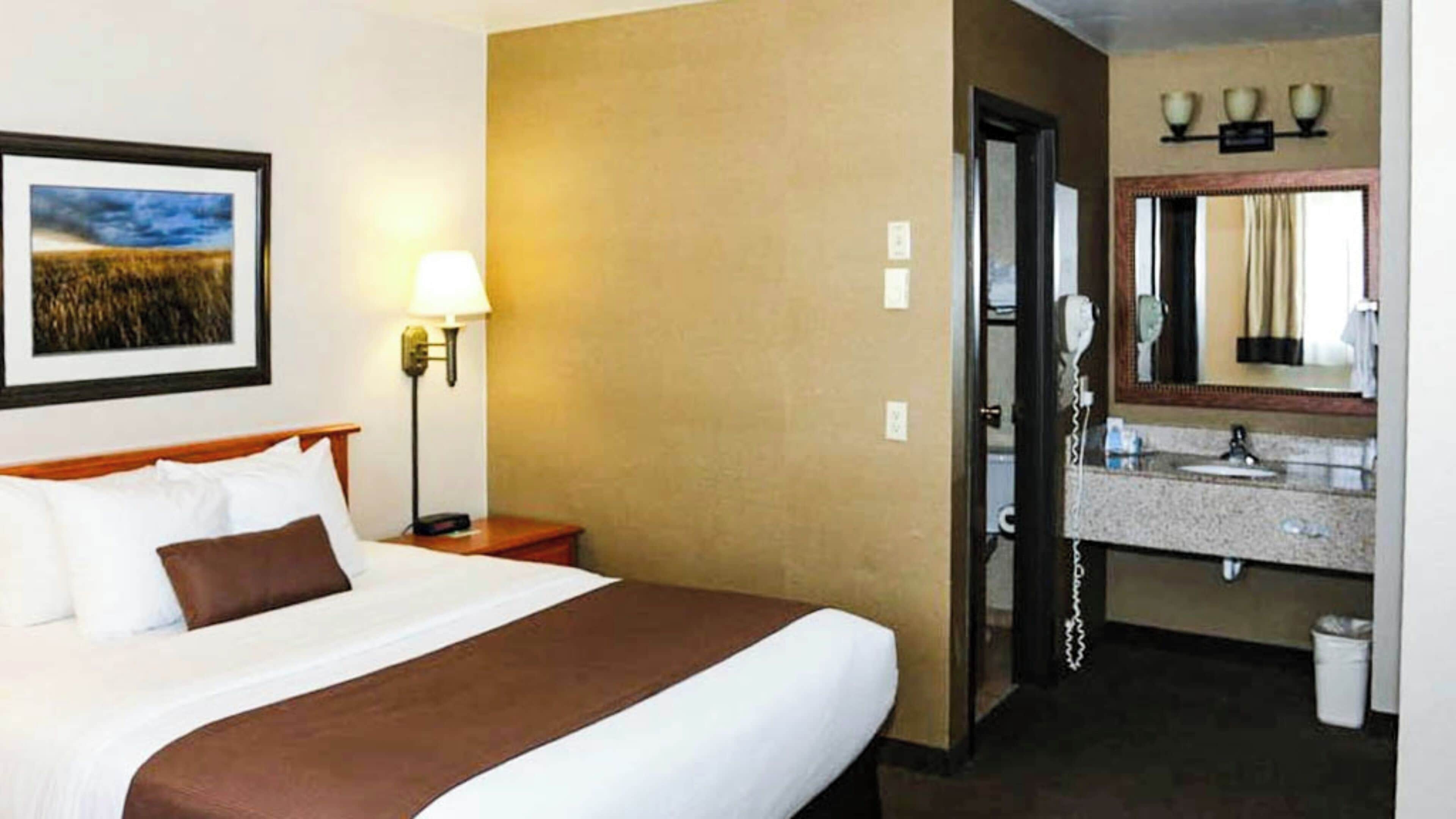 A hotel room with a bed, a bedside lamp, a framed picture on the wall, a bathroom with a sink and mirror, and a telephone.