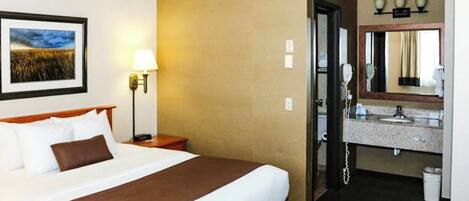 A hotel room with a bed, a bedside lamp, a framed picture on the wall, a bathroom with a sink and mirror, and a telephone.