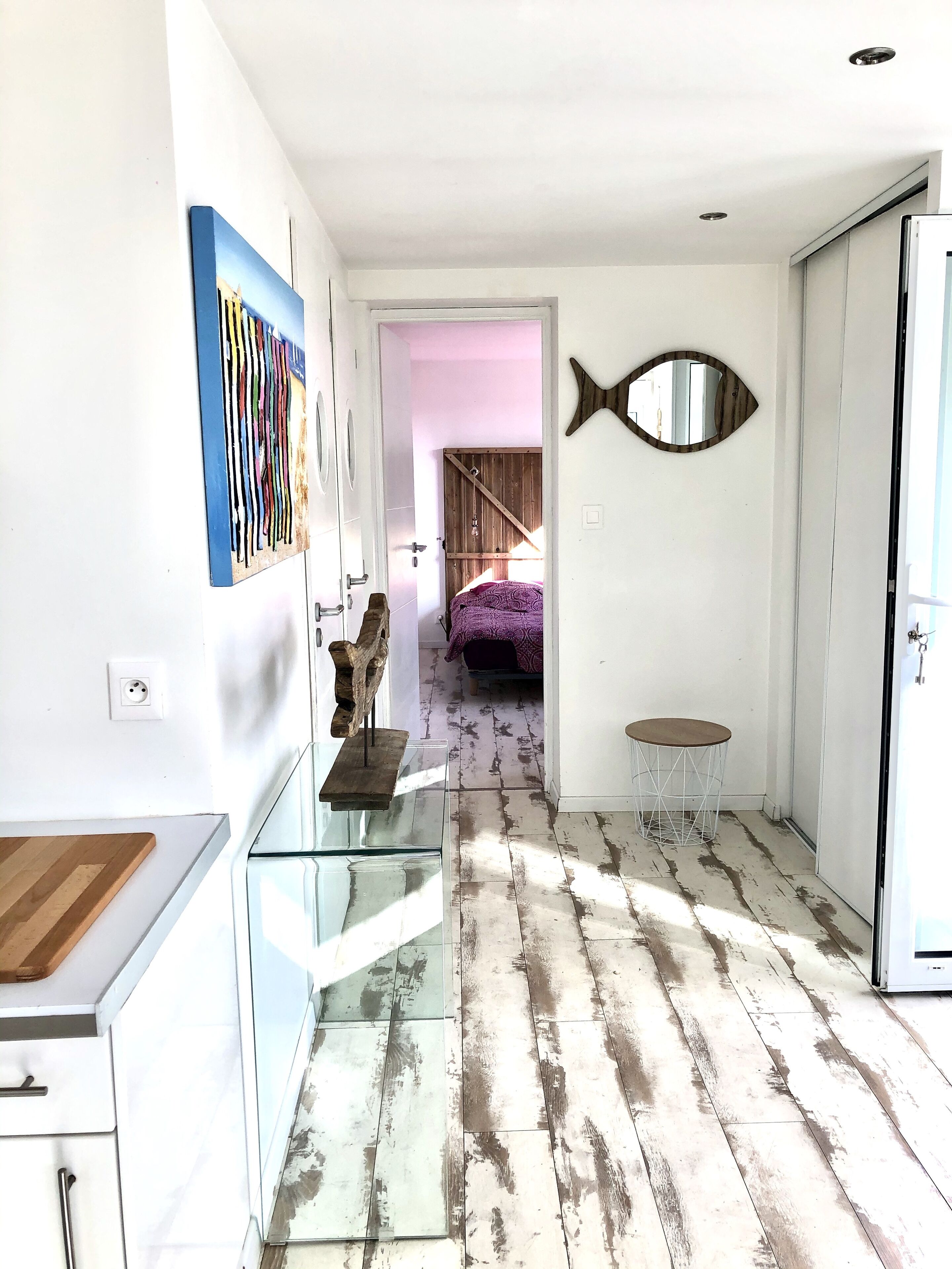 Un pasillo con una mesa de vidrio, un banco de madera, un espejo con forma de pez y una cama al fondo.