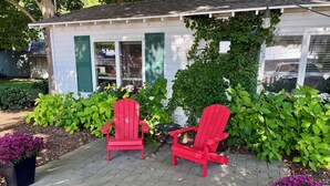 Two red Adirondack chairs in front of a house with green shutters and a garden with various plants and flowers.