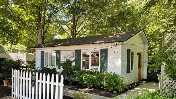 A small, single-story cottage with a white exterior and green shutters, surrounded by a white picket fence and various plants.