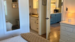A bedroom with a bed, a bedside table, a mirror, and a view into a connected bathroom and kitchen area.