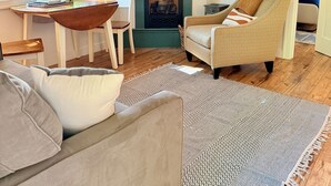 A living room with a fireplace, a TV mounted above it, and a window with blinds. There are two chairs, a sofa, and a patterned rug on the wooden floor.