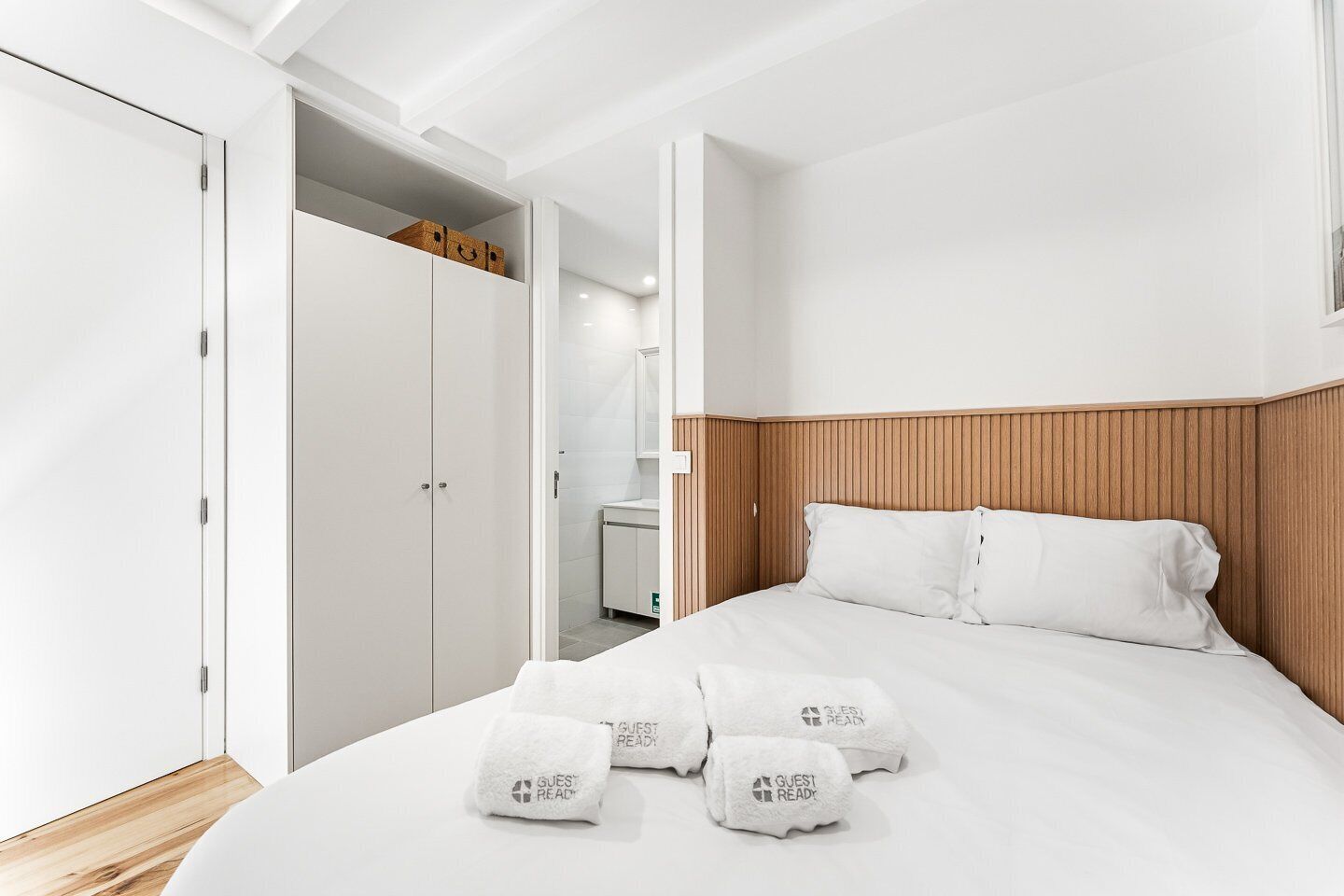 Una camera d'albergo con una testiera in legno, un letto con lenzuola bianche, un armadio con ante bianche e un bagno visibile sullo sfondo.