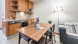 A compact kitchen with wooden cabinets, a Bosch dishwasher, and a microwave. There's a wooden dining table with chairs, a sofa, and a floor lamp.