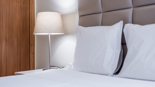 A neatly made bed with white linens, a modern bedside lamp, and a padded headboard.