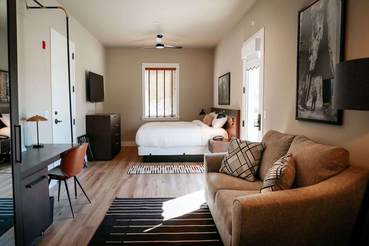 A modern hotel room with a bed, a desk, a chair, a sofa, a ceiling fan, and a window with blinds.