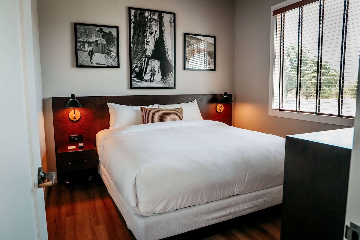 A bedroom with a large bed, two bedside tables with lamps, a window with blinds, and two framed pictures on the wall.