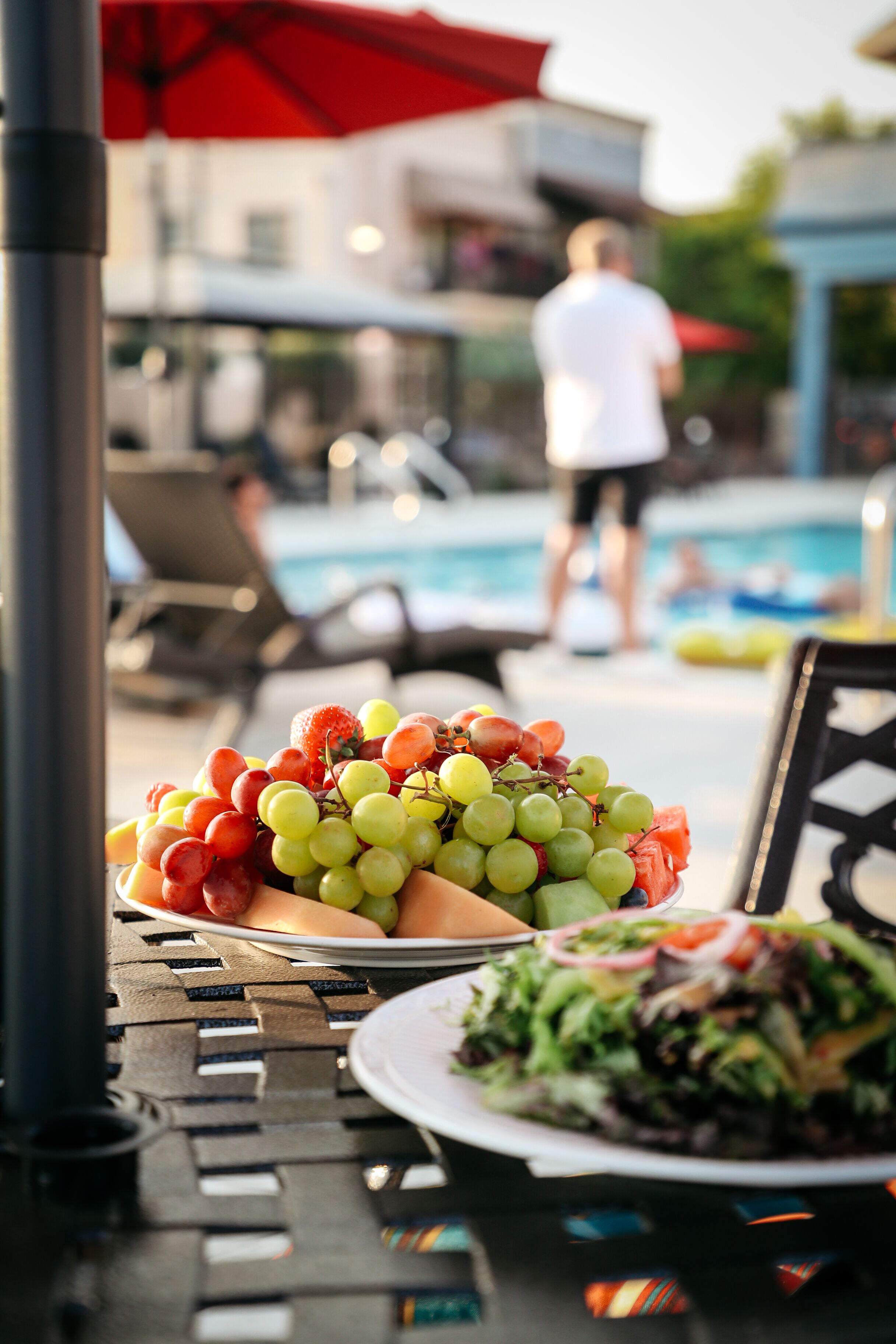 A dining table with a plate of mixed fruits and a salad, a red umbrella, and a pool in the background.