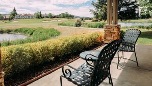 Une terrasse couverte avec deux chaises en métal et une table, donnant sur un terrain de golf et un plan d’eau.