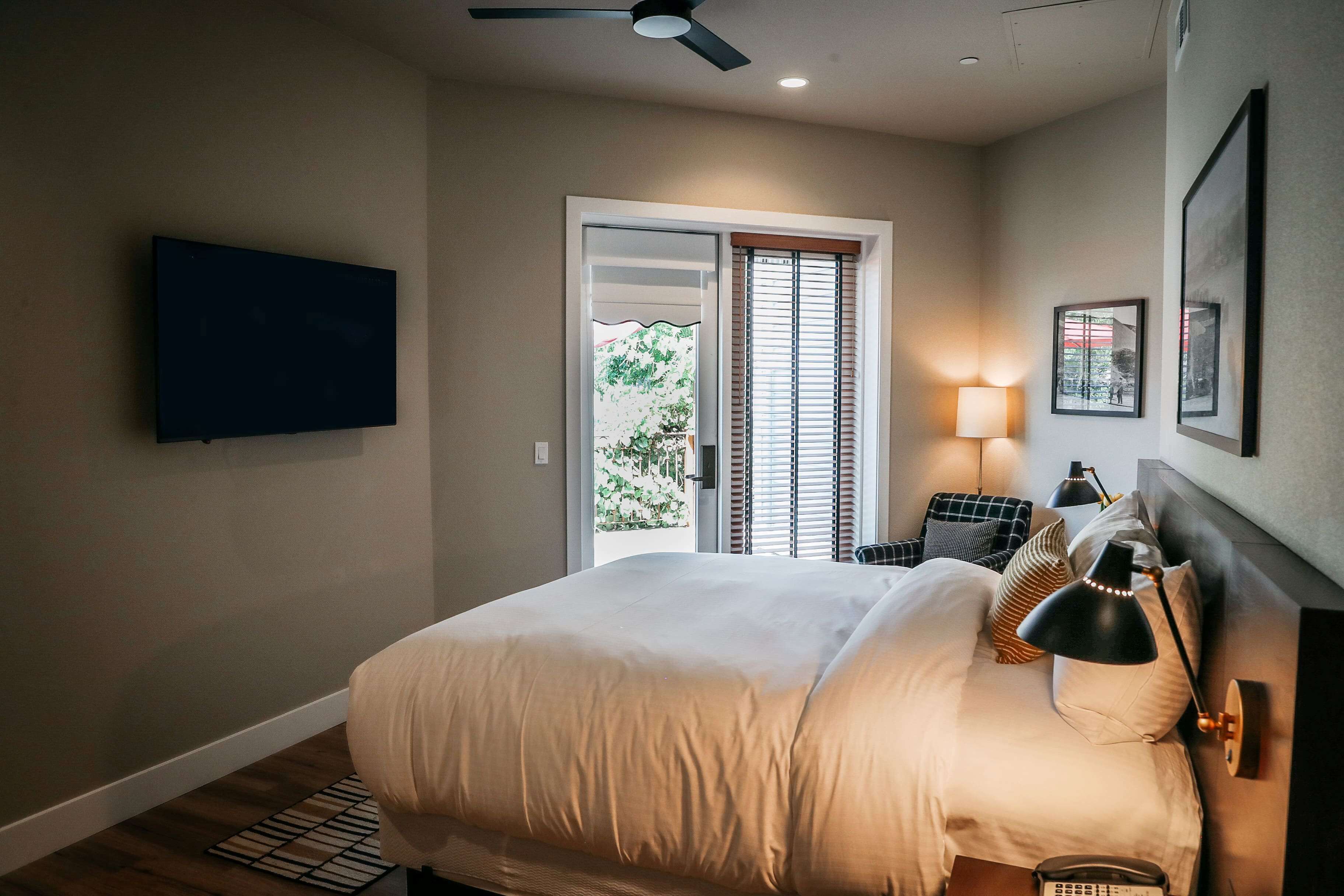 A bedroom with a bed, a ceiling fan, a TV mounted on the wall, a bedside table with a lamp, and a window with blinds.