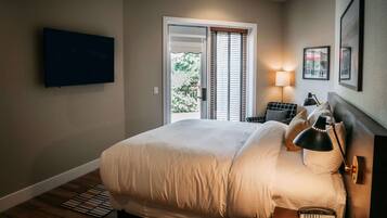 A bedroom with a bed, a ceiling fan, a TV mounted on the wall, a bedside table with a lamp, and a window with blinds.