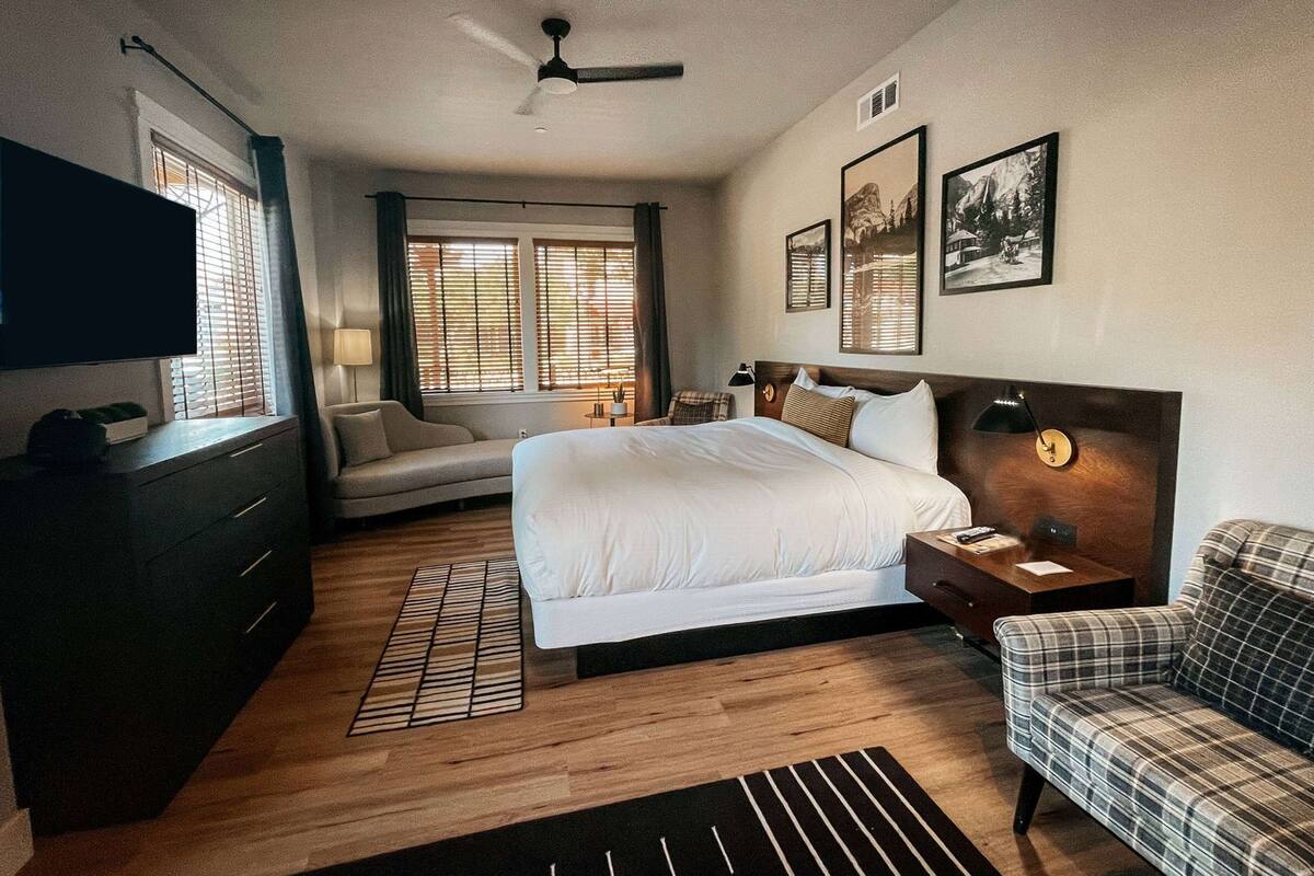 A hotel room with a large bed, a desk, a chair, a TV, and a window with blinds.