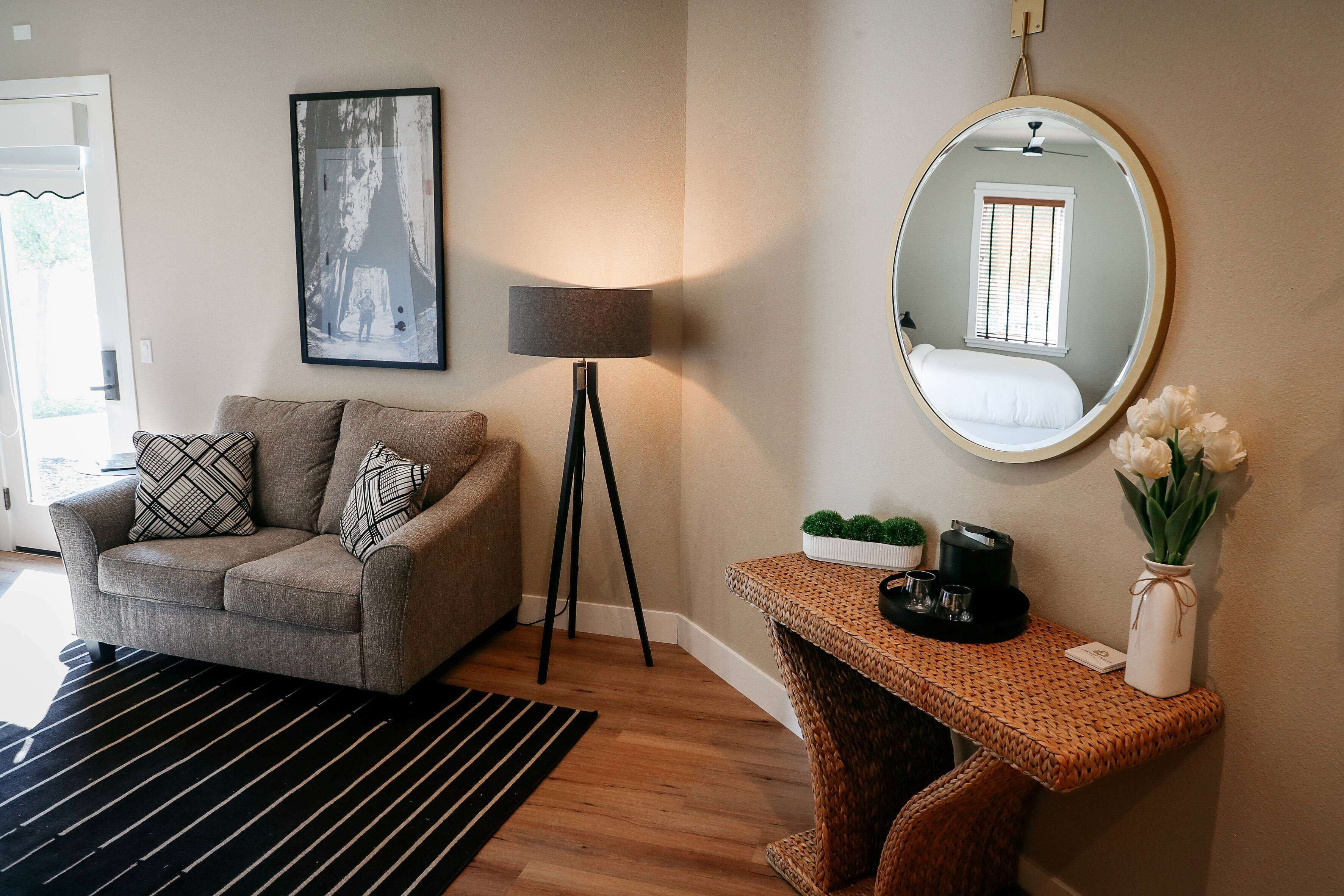 A living room with a sofa, a floor lamp, a round mirror, a small table with a vase of flowers, and a framed picture on the wall.