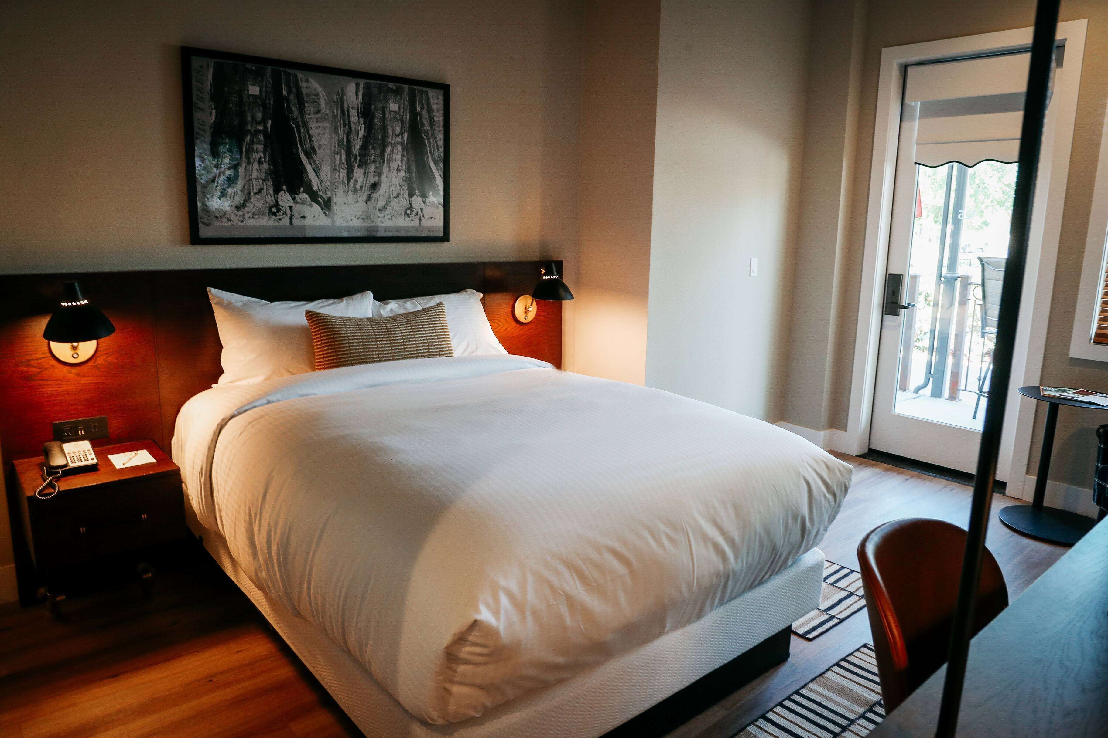 Quarto de hotel com uma cama grande, luminárias de cabeceira, mesa de cabeceira com telefone, pintura na parede e vista para o exterior através de uma porta.