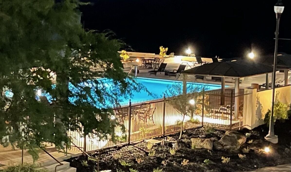 A nighttime view of a swimming pool area with a fenced deck, lounge chairs, and a covered seating area.