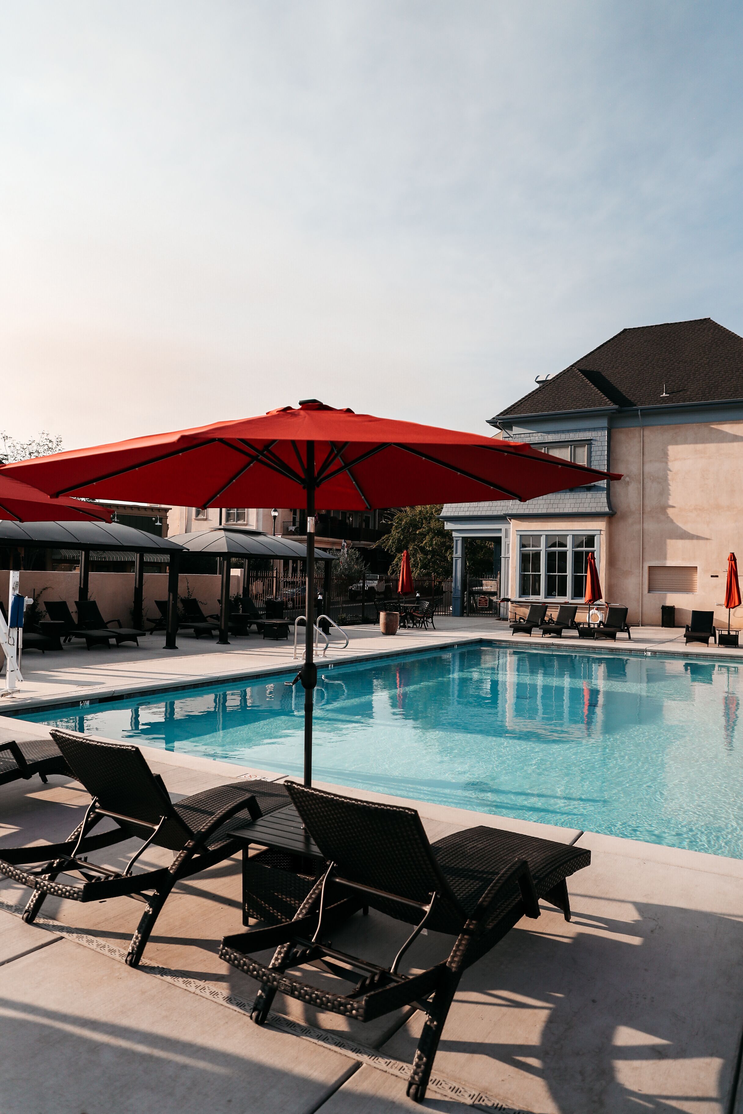 A swimming pool with lounge chairs and umbrellas, a covered seating area, and a building in the background.