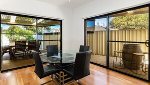 A dining area with a glass table and chairs, large windows, and a view of a backyard with a wooden fence and outdoor furniture. - Swainson on Lexington (Henley Beach South)