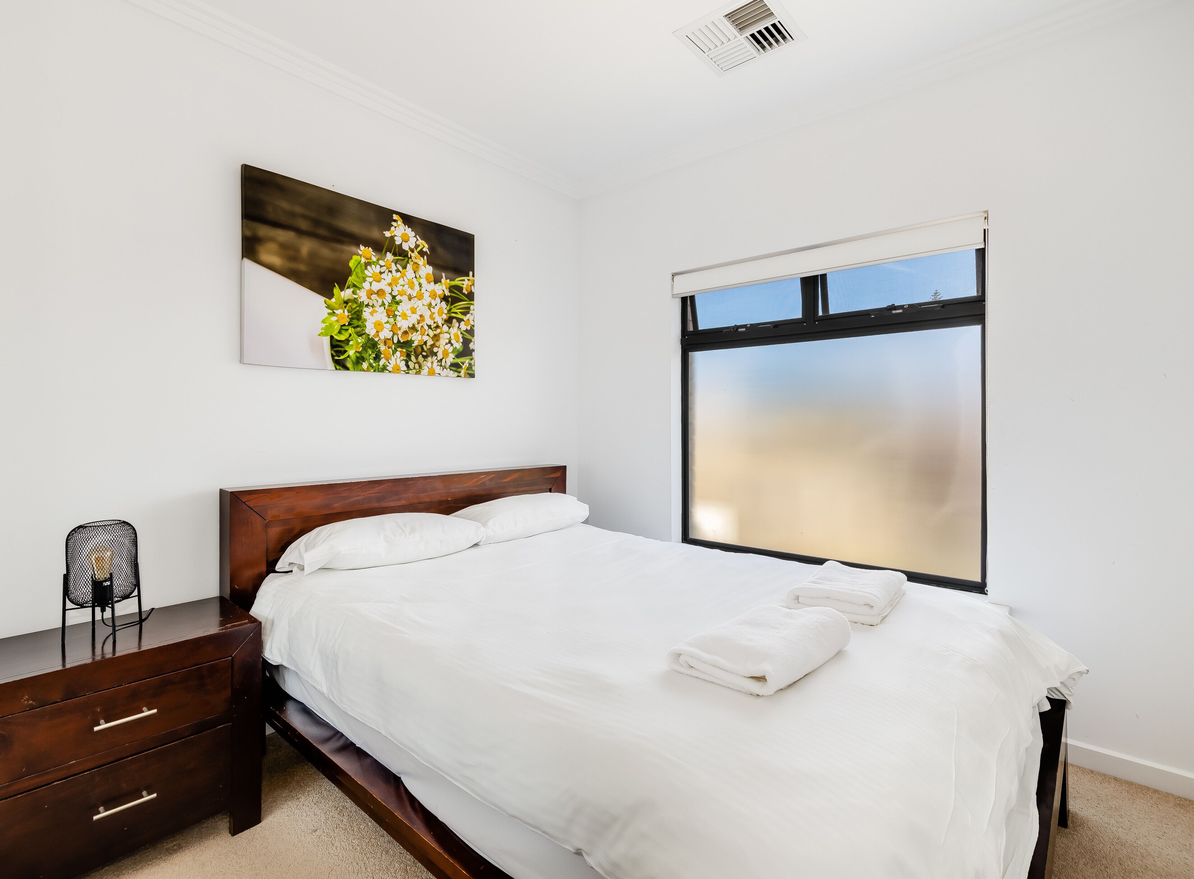 A bedroom with a wooden bed, white bedding, a nightstand, a chair, a wall-mounted picture, and a large window.