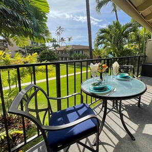 Une table ronde en verre, une chaise avec un coussin bleu, un vase en verre rempli de fleurs et un panier de fruits sont disposés sur un balcon d’où l’on peut admirer une pelouse et des palmiers.