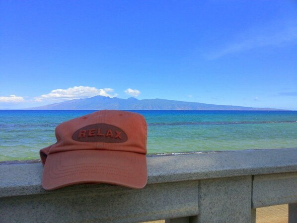 Une casquette rouge avec le mot « RELAX » inscrit dessus, posée sur une rambarde surplombant un plan d’eau et une chaîne de montagnes.