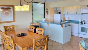 A dining area with a wooden table and chairs, a kitchen with white appliances, and a window with blinds.