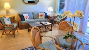 A living room with a sofa, two armchairs, a coffee table, and a glass-top table with chairs. There is a mirror on the wall, a lamp on a side table, and a window with curtains.