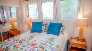Un dormitorio con colcha estampada, dos almohadas azules, una lámpara de noche y una mesita con un control remoto.