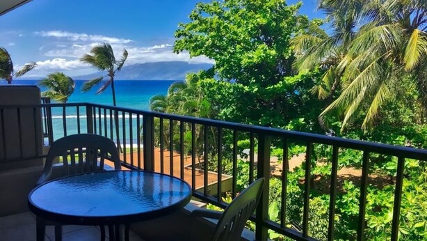 Un balcon avec une table ronde et des chaises, donnant sur un paysage tropical avec des palmiers et une vue sur l’océan.