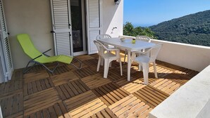 Une terrasse en bois avec une table et des chaises, un fauteuil de jardin vert et une vue sur une colline boisée.
