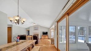 A spacious living room with a wooden dining table, a fireplace, a television, and large windows. - Houghton Lake Getaway w/ Pontoon (Houghton Lake)