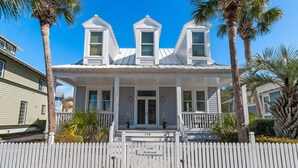 A white colonial-style house with a picket fence, numbered 116, and palm trees in the background.