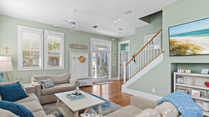 A spacious living room with a staircase, a view of the beach, and a sign that reads "BEACH HOUSE".