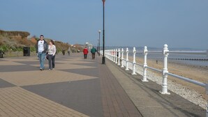 En asfalteret promenade med et hvidt gelænder, en klar himmel og et roligt hav i baggrunden.