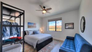 A bedroom with a bed, a blue sofa, a ceiling fan, a window with a view, and a wall-mounted shelf with clothes. - Luxury twin home.  Beautiful views. Private hot tub. Covered outside patio.  (Moab)