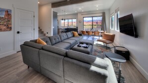 A modern living room with a sectional sofa, a dining area with a table and chairs, a ceiling fan, and large windows offering a view of the desert. - Luxury twin home.  Beautiful views. Private hot tub. Covered outside patio.  (Moab)