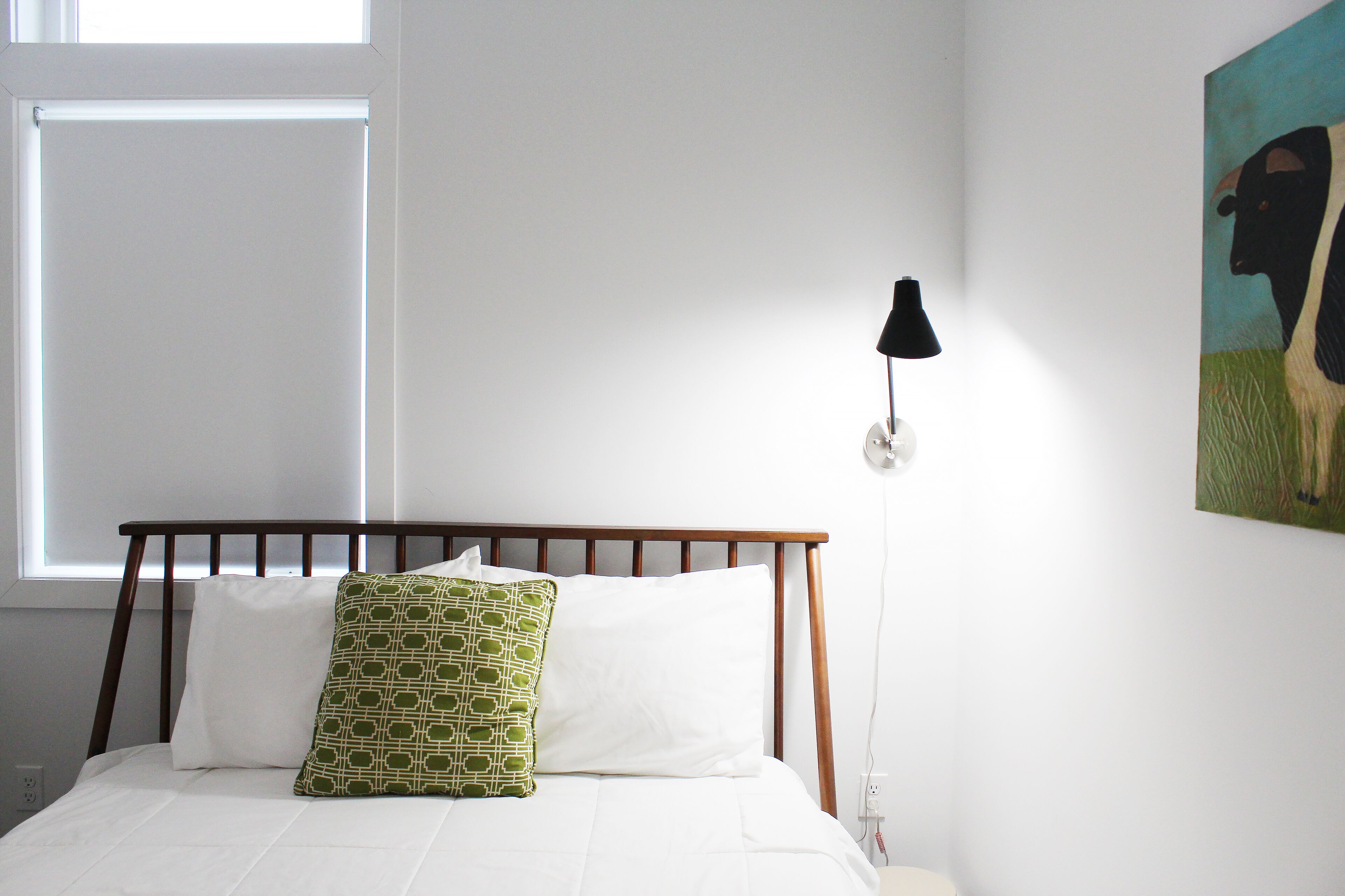 Een bed met witte beddengoed en een kussen met groen patroon, een houten hoofdbord, een zwarte lamp en een schilderij van een koe aan de muur.