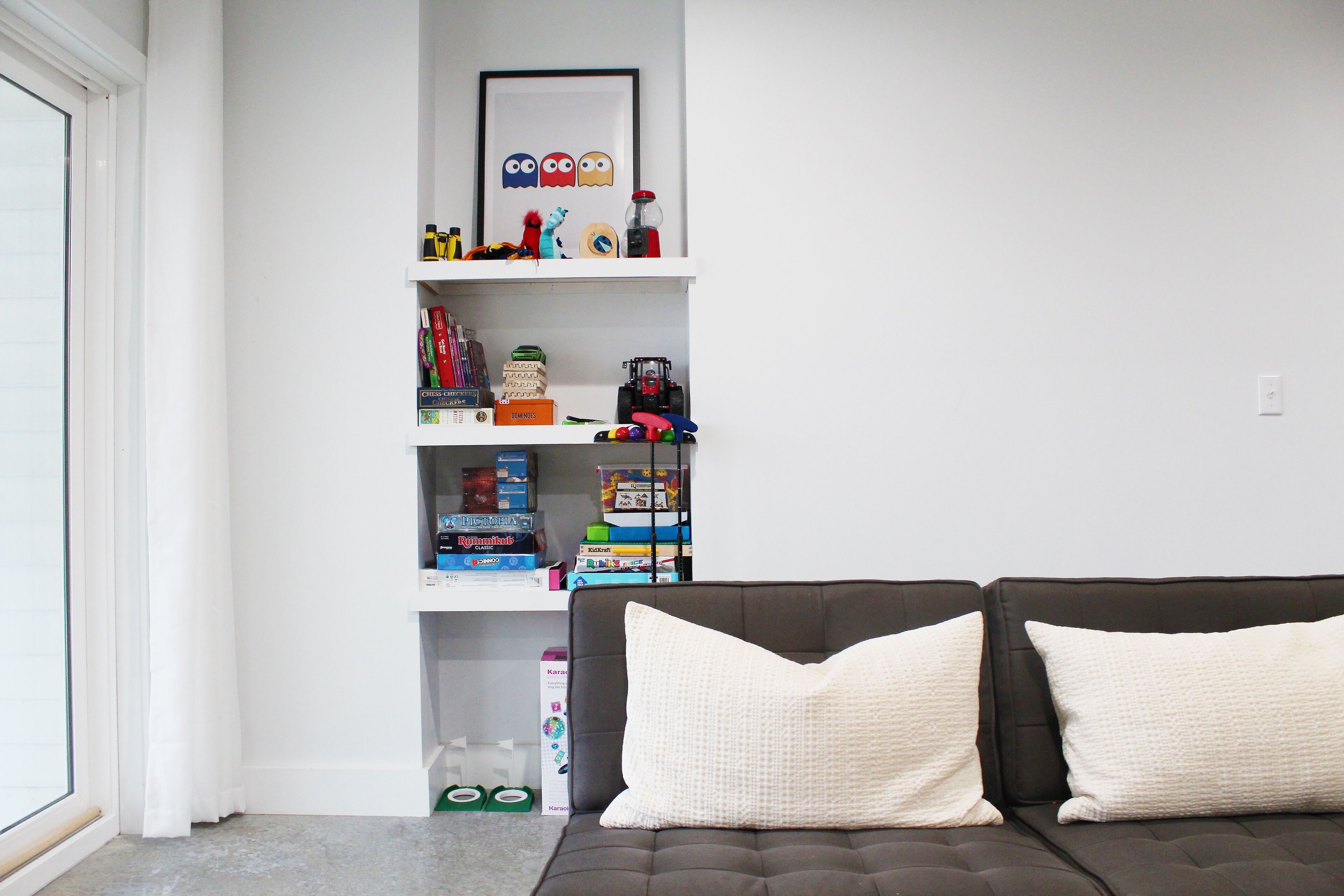 A living room with a dark brown sofa and two white pillows, a white bookshelf with various items, and a window with white curtains.