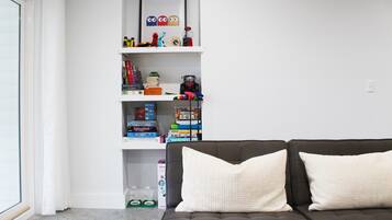 A living room with a dark brown sofa and two white pillows, a white bookshelf with various items, and a window with white curtains.