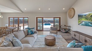 A spacious living room with a large sofa, a dining table, and a view of a pool and mountains through large windows. - 3 Bedroom Private Home at Waikoloa Beach with Pool (Waikoloa)