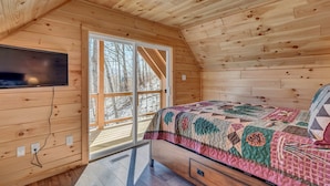 A cozy cabin room with a wooden ceiling, a large window, a bed with a colorful quilt, and a flat-screen TV mounted on the wall. - Cozy, Luxurious Fox Den in the Shenandoah Valley (Luray)