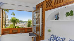 A bedroom with a bed, a small table with a laptop, a window with wooden shutters, and a view of a beach and buildings. - Apartamento Carabela 51 (Puerto de Alcudia)