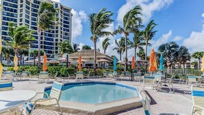 A pool area with lounge chairs and umbrellas, surrounded by palm trees and a building in the background. - Beautiful Hutchinson Island Condo - Steps to Ocean (Fort Pierce)